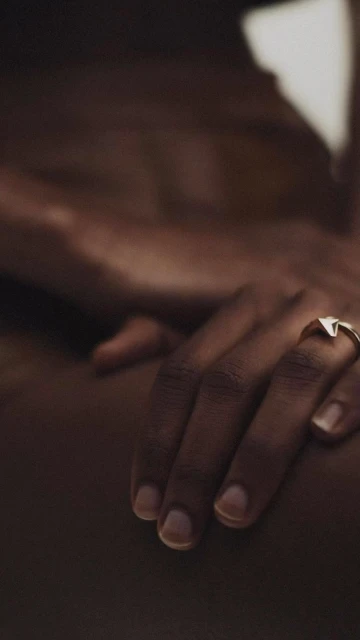 Close-up studio shot of a dark-skinned person's hands with a gold ring, bathed in warm light, showcasing minimal nude skin.