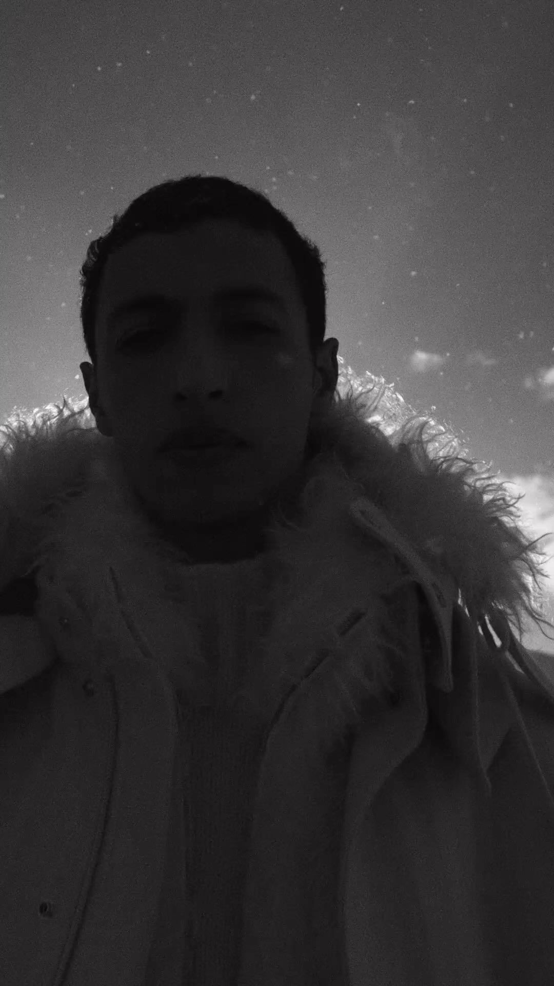 Low-angle monochrome portrait of a man in a textured fur-collar coat against a cloudy sky.