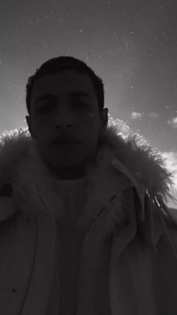 Low-angle monochrome portrait of a man in a textured fur-collar coat against a cloudy sky.