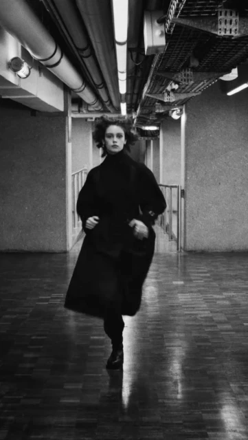 Black and white shot of a woman in a dark coat running in a corridor with lights and pipes on the ceiling, creating a dynamic, stylized image.