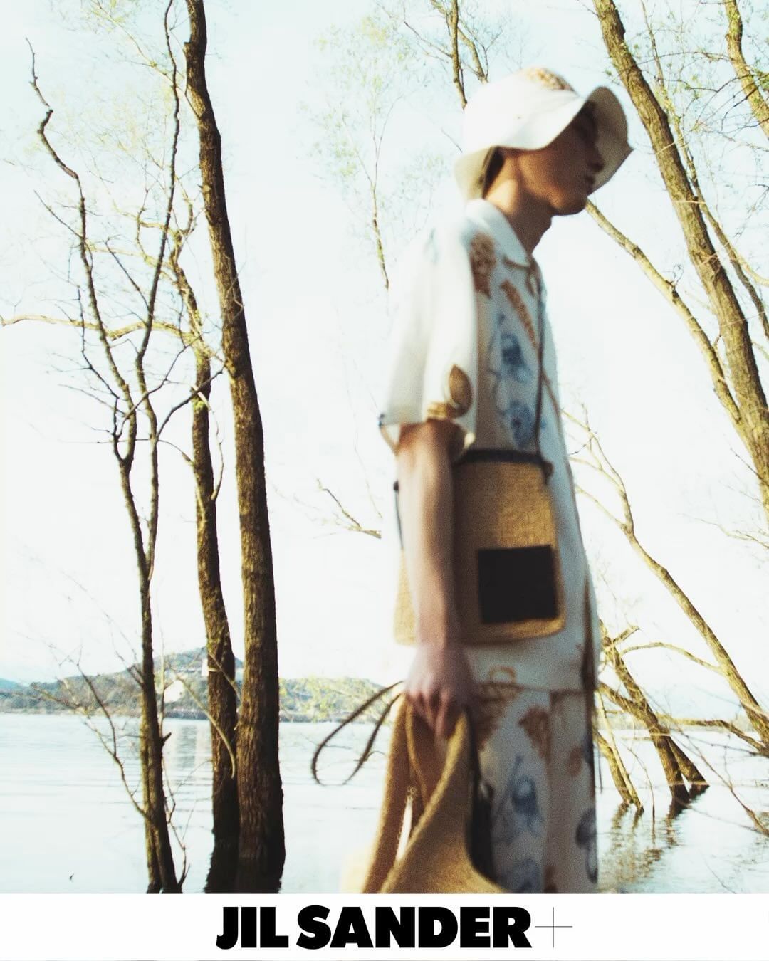 Fashion shot of a person wearing a patterned outfit with a matching hat and shoulder bag, posing in front of a lake and trees.