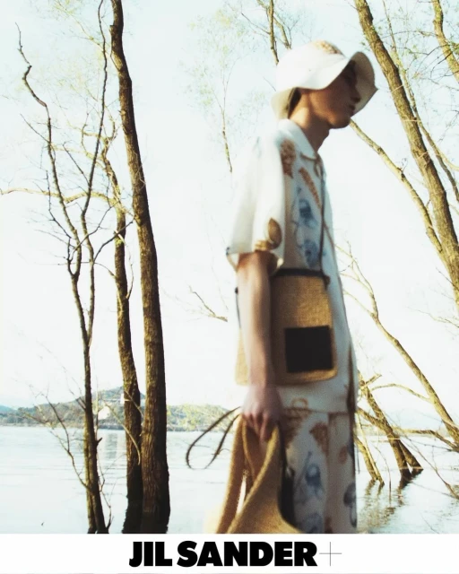 Fashion shot of a person wearing a patterned outfit with a matching hat and shoulder bag, posing in front of a lake and trees.