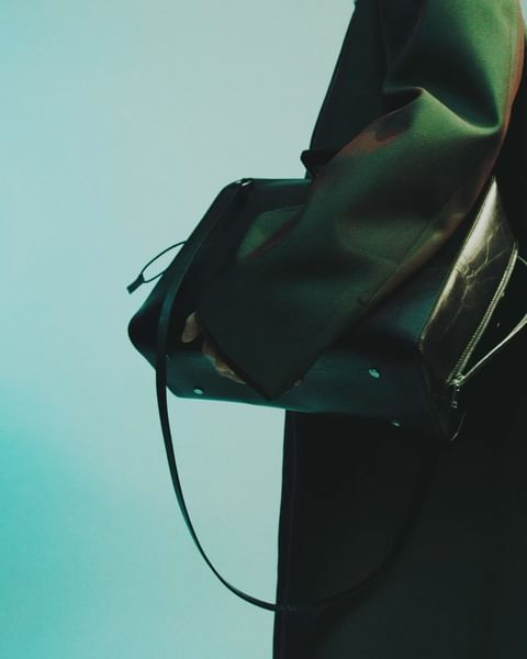 A dark green leather bag with a strap is held by a person wearing a coat, against a turquoise background.