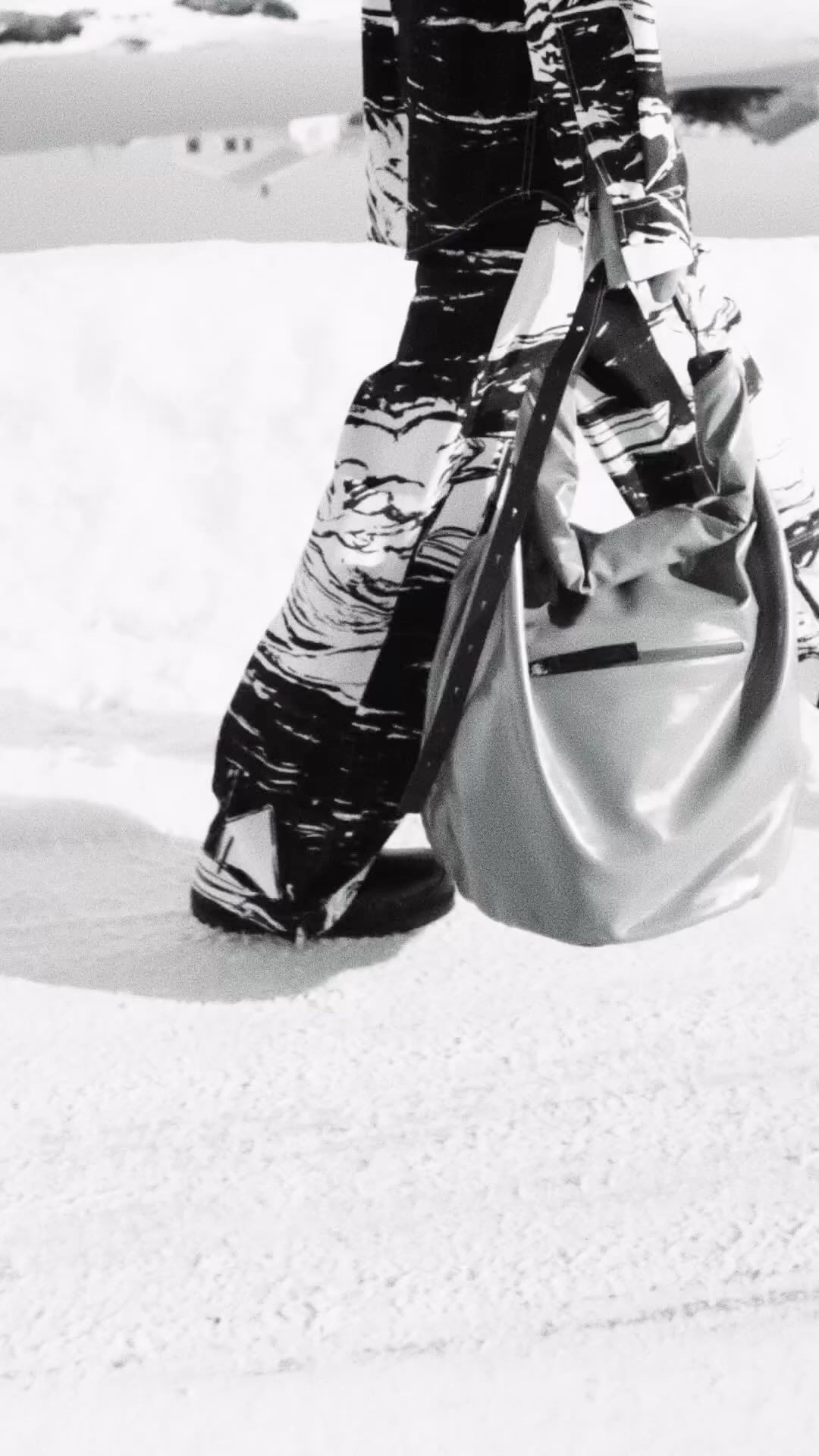 Monochrome fashion photograph showing patterned trousers and a large slouchy shoulder bag outdoors in snow