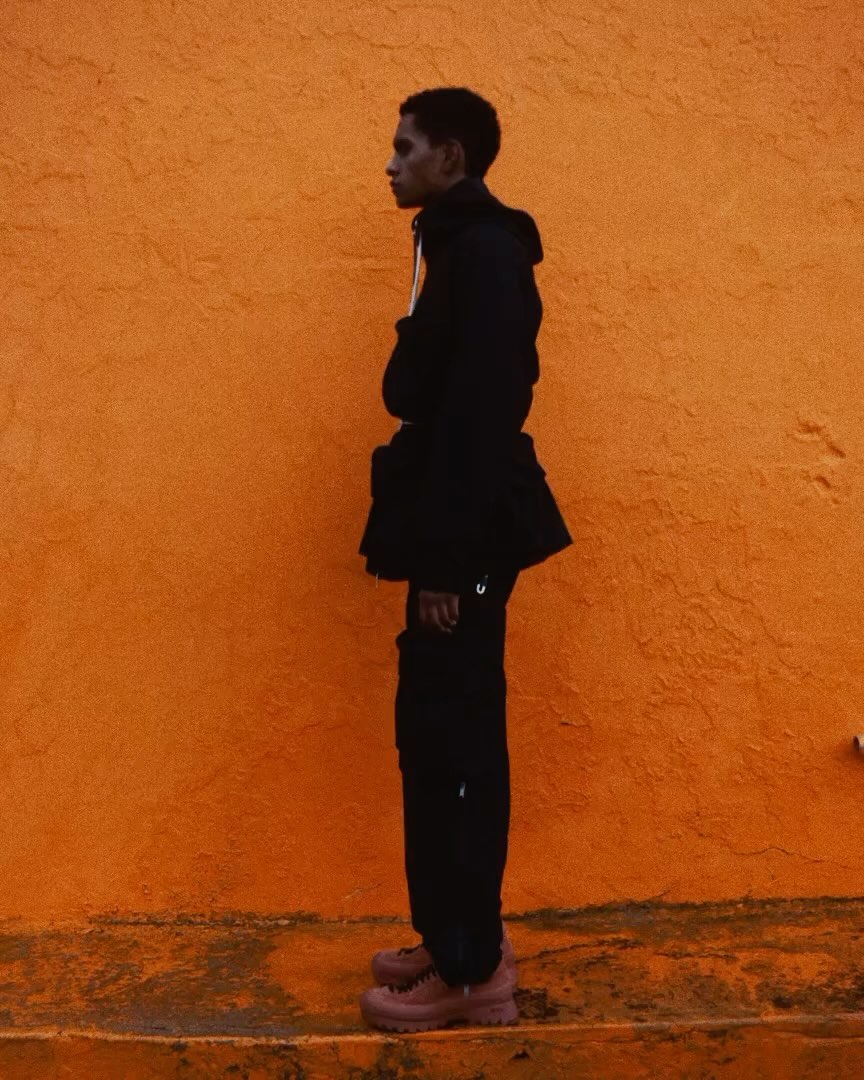 A young adult stands in profile against an orange wall, wearing a dark hooded jacket, dark trousers, and dark pink platform shoes.