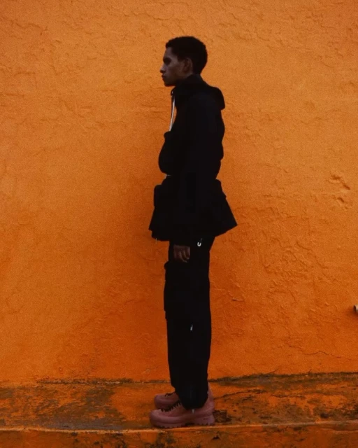 A young adult stands in profile against an orange wall, wearing a dark hooded jacket, dark trousers, and dark pink platform shoes.