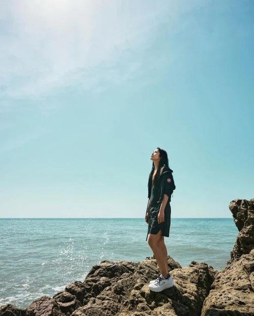 Woman in dark romper and white sneakers poses on rocky outcrop with ocean and blue sky in background on a bright sunny day.