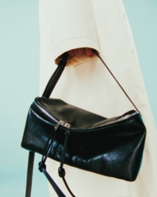 Black leather shoulder bag with a silver zipper is held by a person wearing a cream-colored coat against a light teal backdrop.