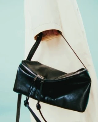 Black leather shoulder bag with a silver zipper is held by a person wearing a cream-colored coat against a light teal backdrop.