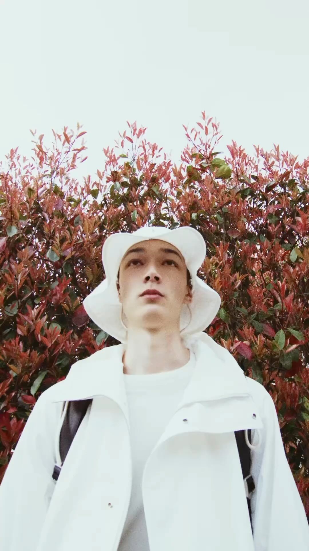 Young adult male poses in a white outfit with a matching hat against a backdrop of red leaves and a bright, overcast sky.