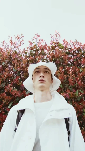 Young adult male poses in a white outfit with a matching hat against a backdrop of red leaves and a bright, overcast sky.