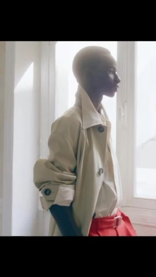 Woman in trench coat stands by a window in a bright indoor setting, looking away. Light neutral and warm color grading.