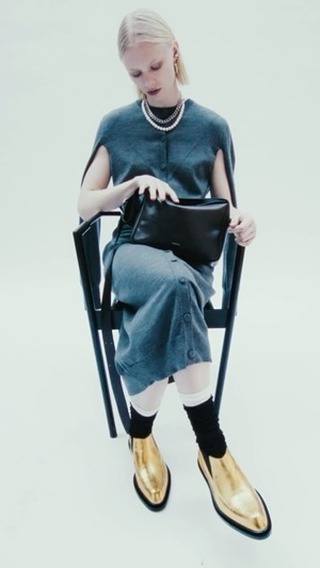 A woman is sitting on a chair indoors, holding a black purse, and wearing a grey sweater dress, white socks, and shiny gold boots.