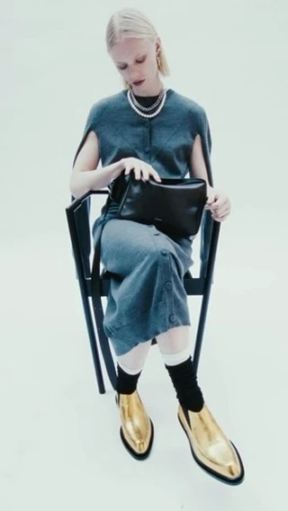 A woman is sitting on a chair indoors, holding a black purse, and wearing a grey sweater dress, white socks, and shiny gold boots.