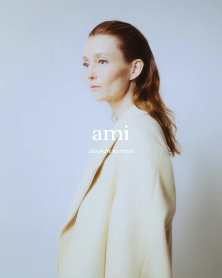 Portrait of a red-haired adult female in a cream-colored coat, posing against a light blue background, with visible 'ami' logo.