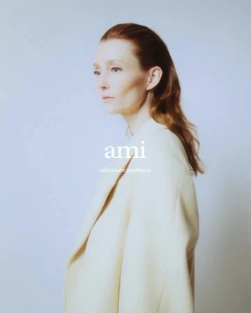 Portrait of a red-haired adult female in a cream-colored coat, posing against a light blue background, with visible 'ami' logo.