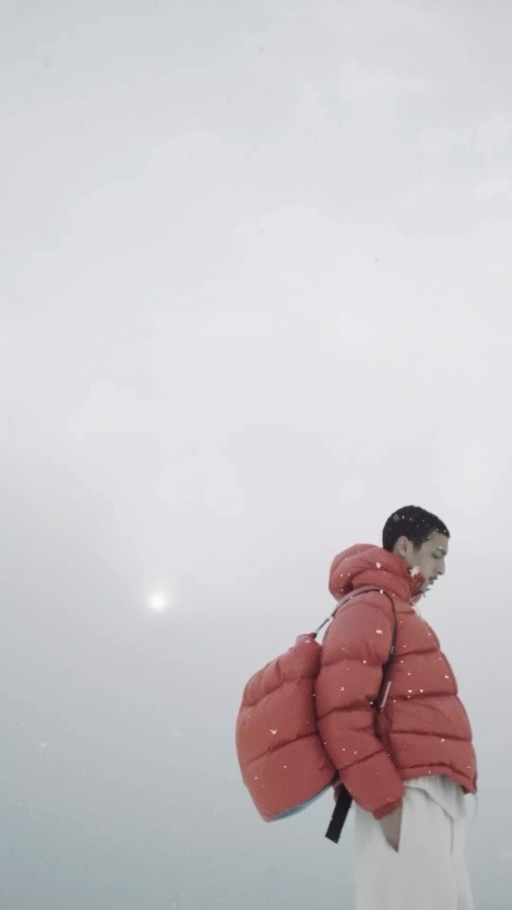 A person walks through a snowy, muted landscape, wearing a puffy red coat and carrying a matching red backpack.