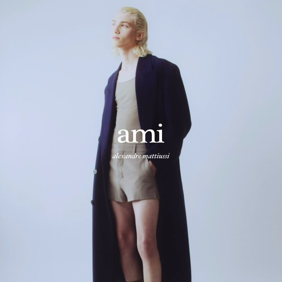 Fashion model in a studio setting wearing a tank top, shorts, and a long coat, posing against a solid light background.