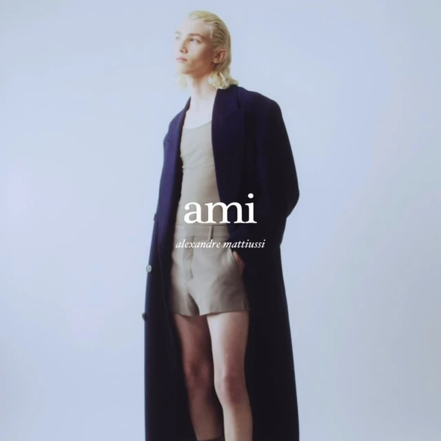 Fashion model in a studio setting wearing a tank top, shorts, and a long coat, posing against a solid light background.