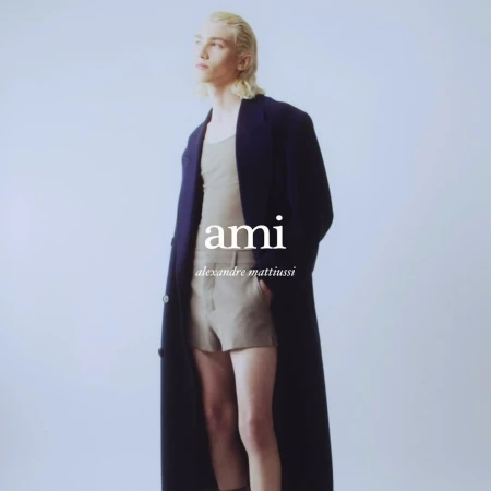 Fashion model in a studio setting wearing a tank top, shorts, and a long coat, posing against a solid light background.