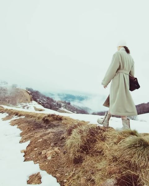 Woman in long coat walking on grassy, snowy hilltop with trees and mountains in the background on a cloudy, overcast day.