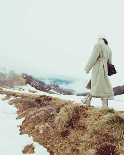 Woman in long coat walking on grassy, snowy hilltop with trees and mountains in the background on a cloudy, overcast day.