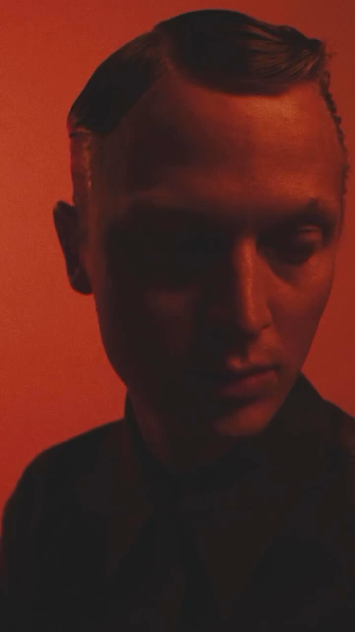 Portrait of a person against an orange backdrop with intense lighting, featuring a serious expression and a dark silhouette.