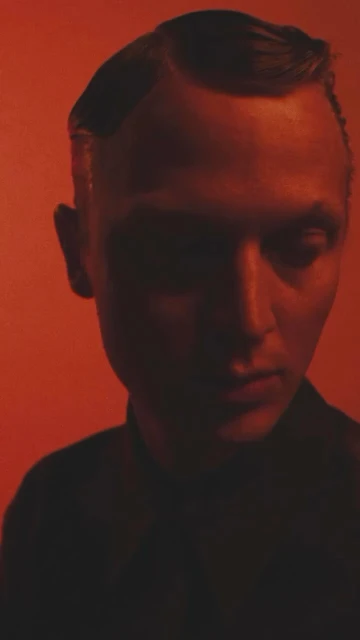 Portrait of a person against an orange backdrop with intense lighting, featuring a serious expression and a dark silhouette.