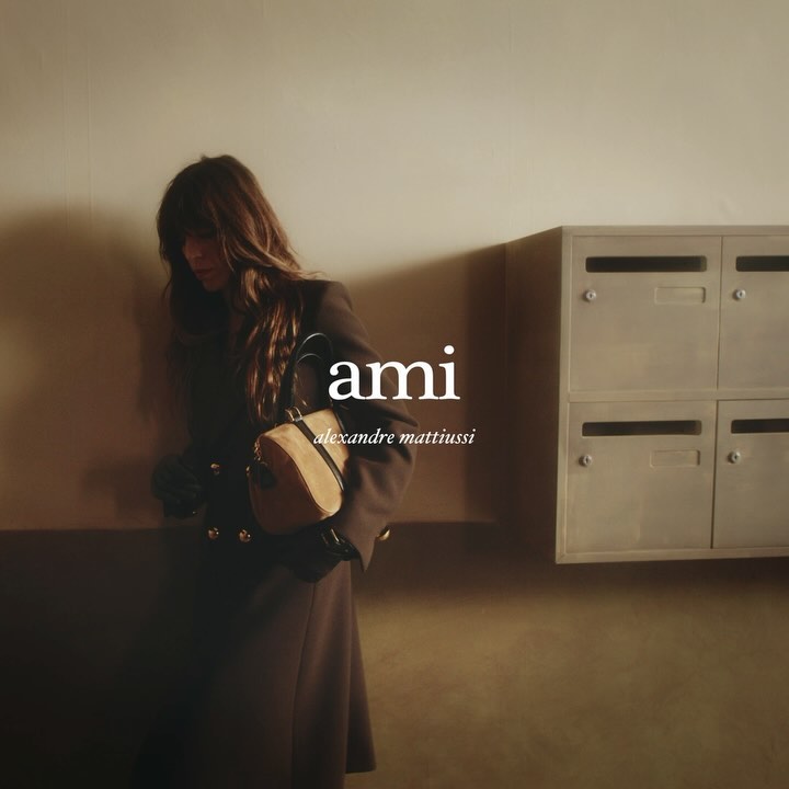 A woman in a brown coat and carrying a beige bag is seen in an indoor setting with metal mailboxes and Ami branding.