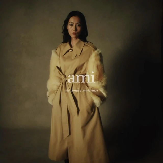Woman in a tan trench coat with fur trim on the sleeves poses against a neutral background. The "ami" logo is visible.