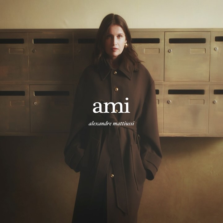 A woman in a brown overcoat stands in front of a wall of metal mailboxes, showcasing the "ami alexandre mattiussi" brand logo.