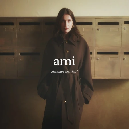 A woman in a brown overcoat stands in front of a wall of metal mailboxes, showcasing the "ami alexandre mattiussi" brand logo.