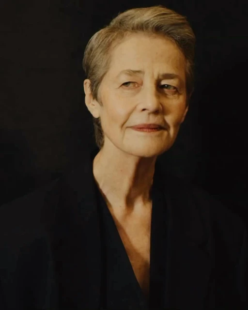 Close-up portrait of an elderly woman with short grey hair wearing a dark blazer against a dark background.