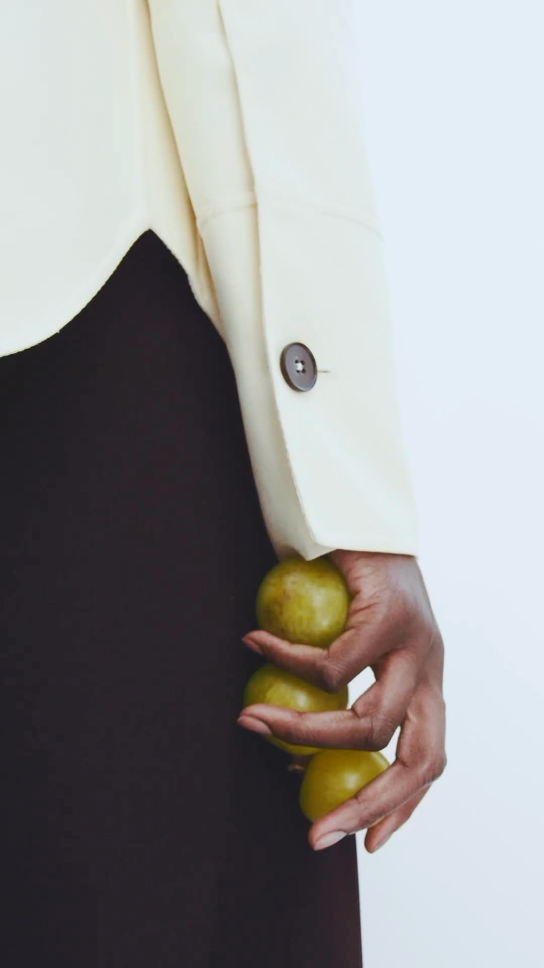 Close-up of a hand holding three green plums against a white background with a light beige jacket and a dark brown skirt.
