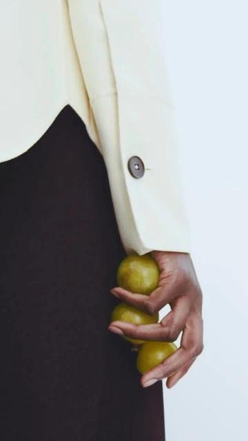 Close-up of a hand holding three green plums against a white background with a light beige jacket and a dark brown skirt.