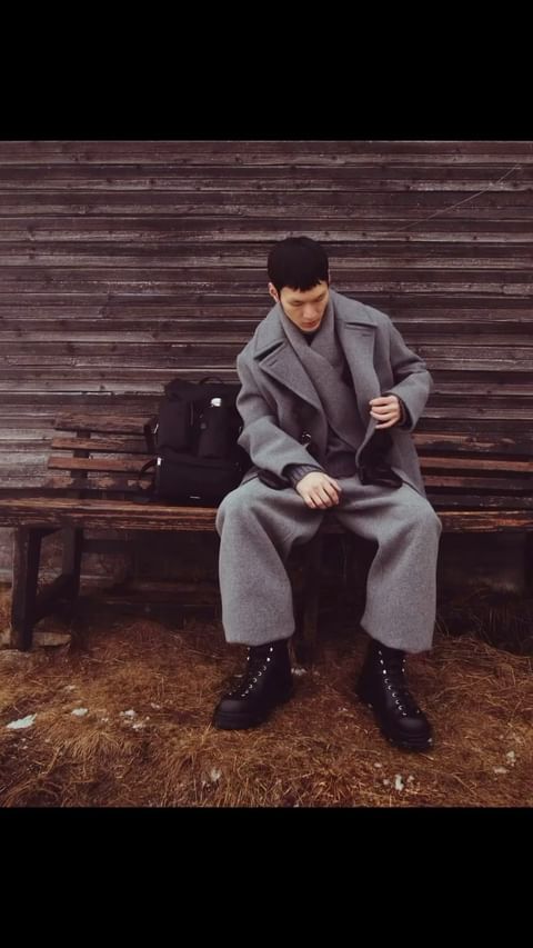 A seated adult male with a backpack poses on a bench in an outdoor setting. He wears a gray coat, pants, and black boots.