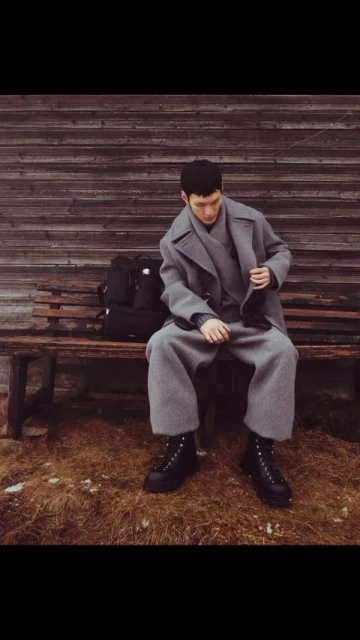 A seated adult male with a backpack poses on a bench in an outdoor setting. He wears a gray coat, pants, and black boots.