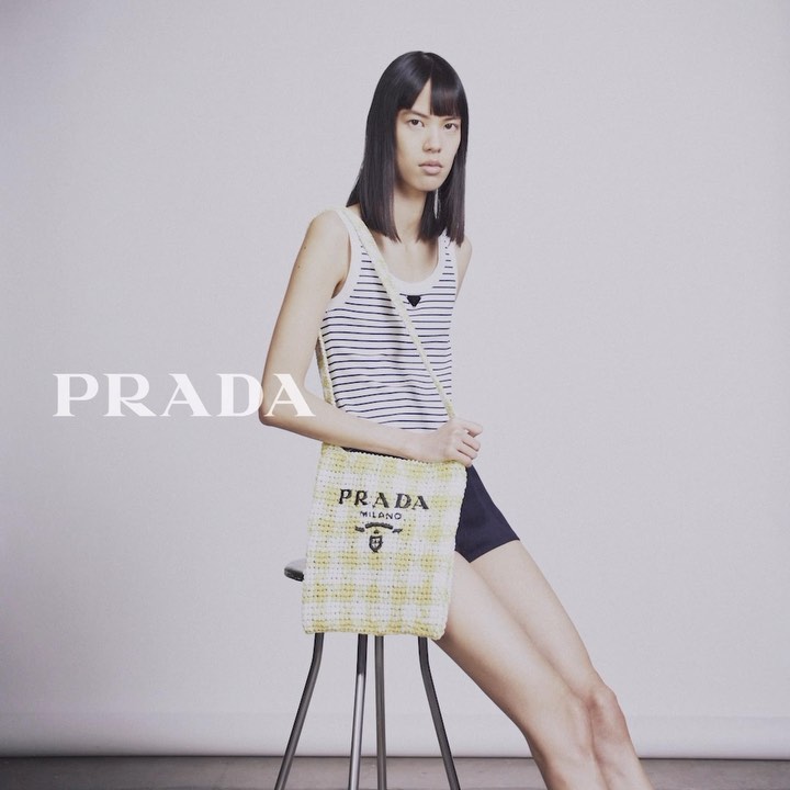 A stylish woman poses in a studio with a Prada handbag, showcasing a striped tank top and black shorts against a neutral backdrop.