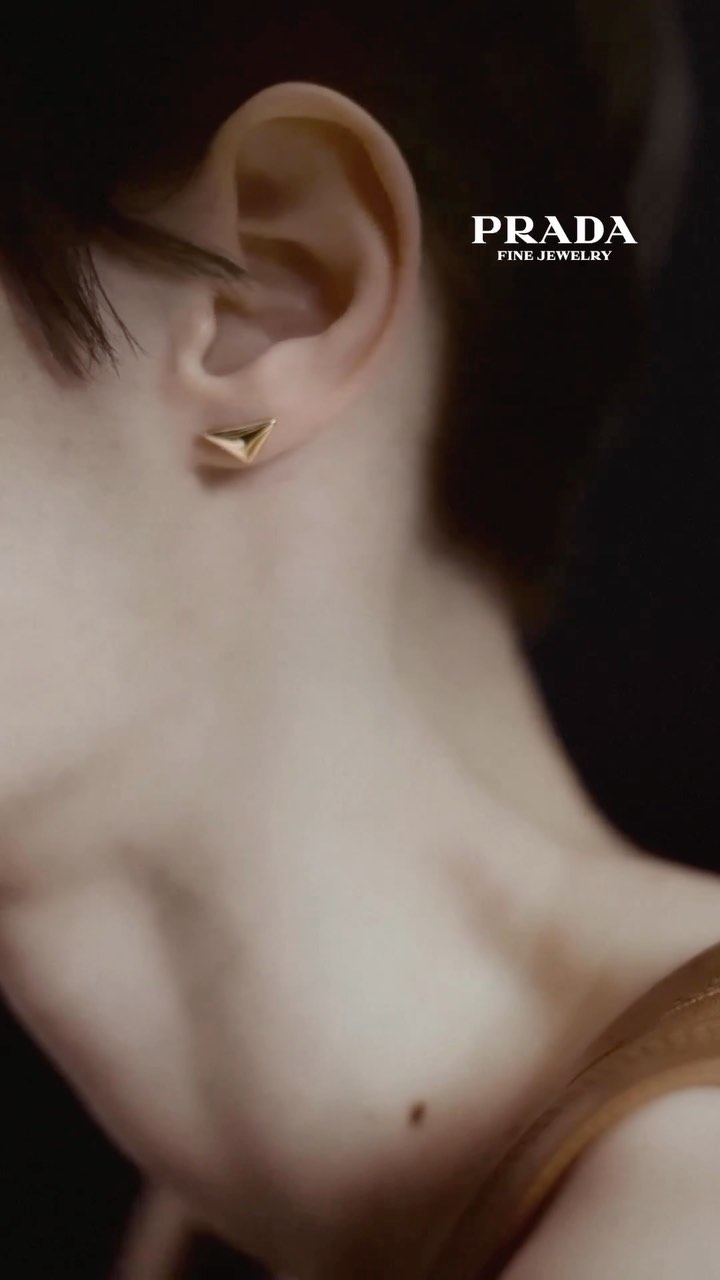 Close-up of a fair-skinned woman's ear with a gold Prada earring, shallow focus on the jewelry against a dark, blurred background.
