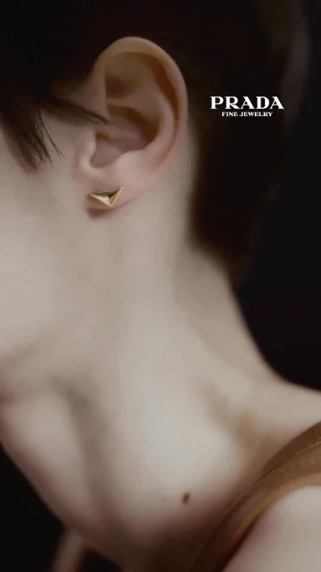 Close-up of a fair-skinned woman's ear with a gold Prada earring, shallow focus on the jewelry against a dark, blurred background.