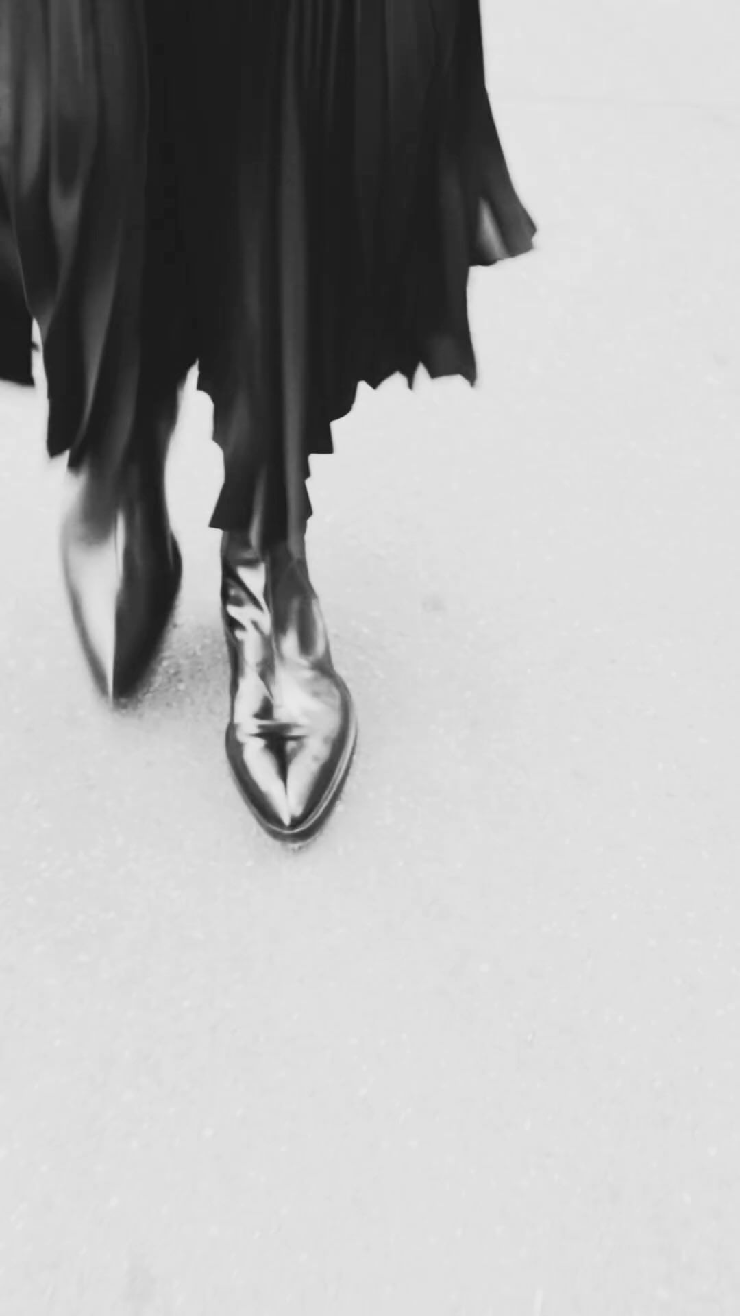 Close-up of metallic, pointed-toe ankle boots and a black, flowing dress on a gray surface, creating a dynamic, monochrome fashion image.