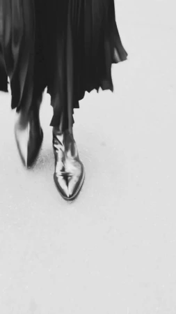 Close-up of metallic, pointed-toe ankle boots and a black, flowing dress on a gray surface, creating a dynamic, monochrome fashion image.