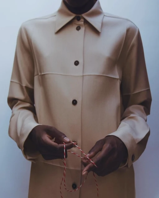 Person in a tan button-down shirt with dark skin tying a red and white string indoors, with a neutral wall backdrop and studio lighting.