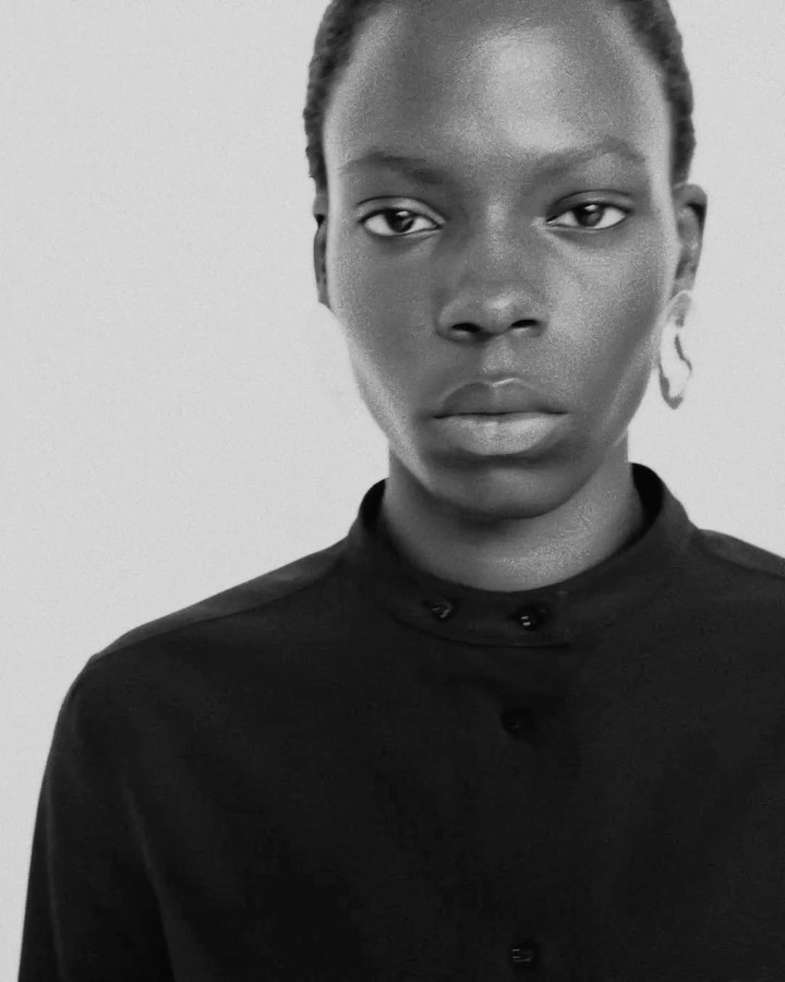 Close-up portrait of a Black female fashion model in a black collared shirt against a white background. Studio lighting highlights her features.