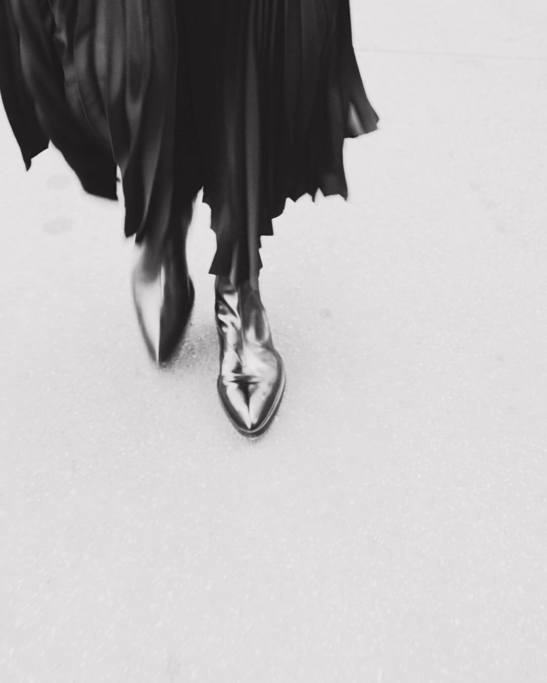 Fashionable black leather ankle boots with a pointed toe under a flowing black dress, captured at a high angle in a monochromatic setting.