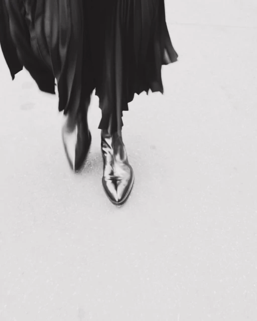 Fashionable black leather ankle boots with a pointed toe under a flowing black dress, captured at a high angle in a monochromatic setting.