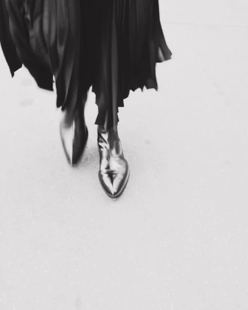 Fashionable black leather ankle boots with a pointed toe under a flowing black dress, captured at a high angle in a monochromatic setting.