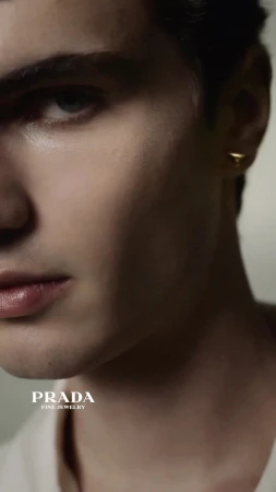Close-up portrait of a young man wearing a gold triangle earring against a neutral background with Prada Fine Jewelry logo visible