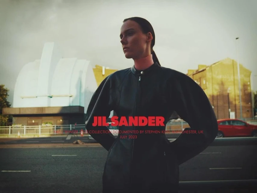 Fashion shot of a woman in a black leather outfit in an urban setting with a domed building in the background. Jil Sander branding.