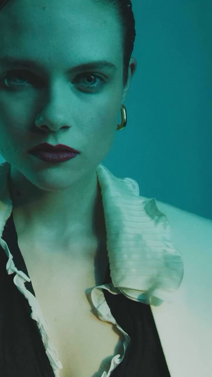Portrait of a young woman with red lipstick and gold earrings, wearing a black top with a white ruffled collar, against a teal background.